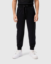 A person stands wearing the Psycho Bunny KIDS WALTER LIGHTWEIGHT SWEATPANT - B0P860D200 in black paired with white sneakers. The drawstring of the lightweight sweatpant is visible at the waist, and the person's hands are relaxed by their sides. The plain light gray background highlights the casual wear perfectly.