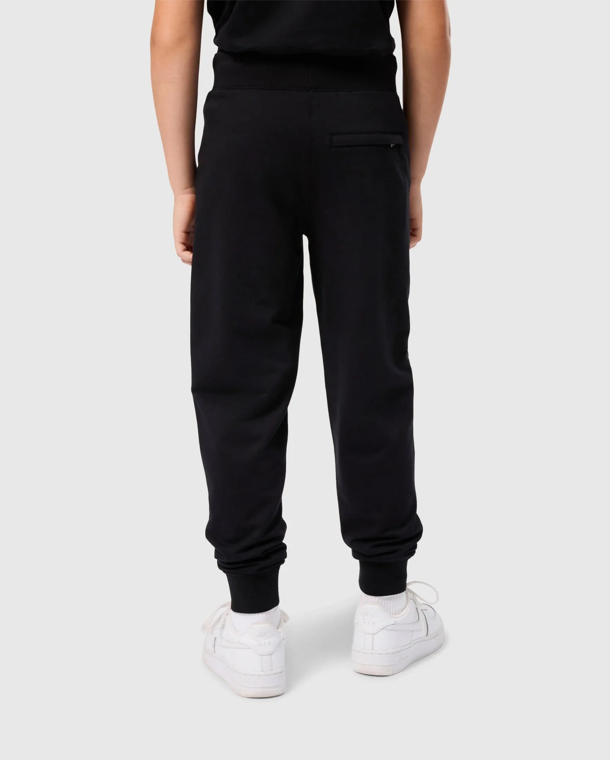 A child is shown wearing black KIDS WALTER LIGHTWEIGHT SWEATPANT - B0P860D200 by Psycho Bunny and white sneakers, from the back. This lightweight sweatpant features a pocket on the upper right side. The plain white background emphasizes the casual wear look.
