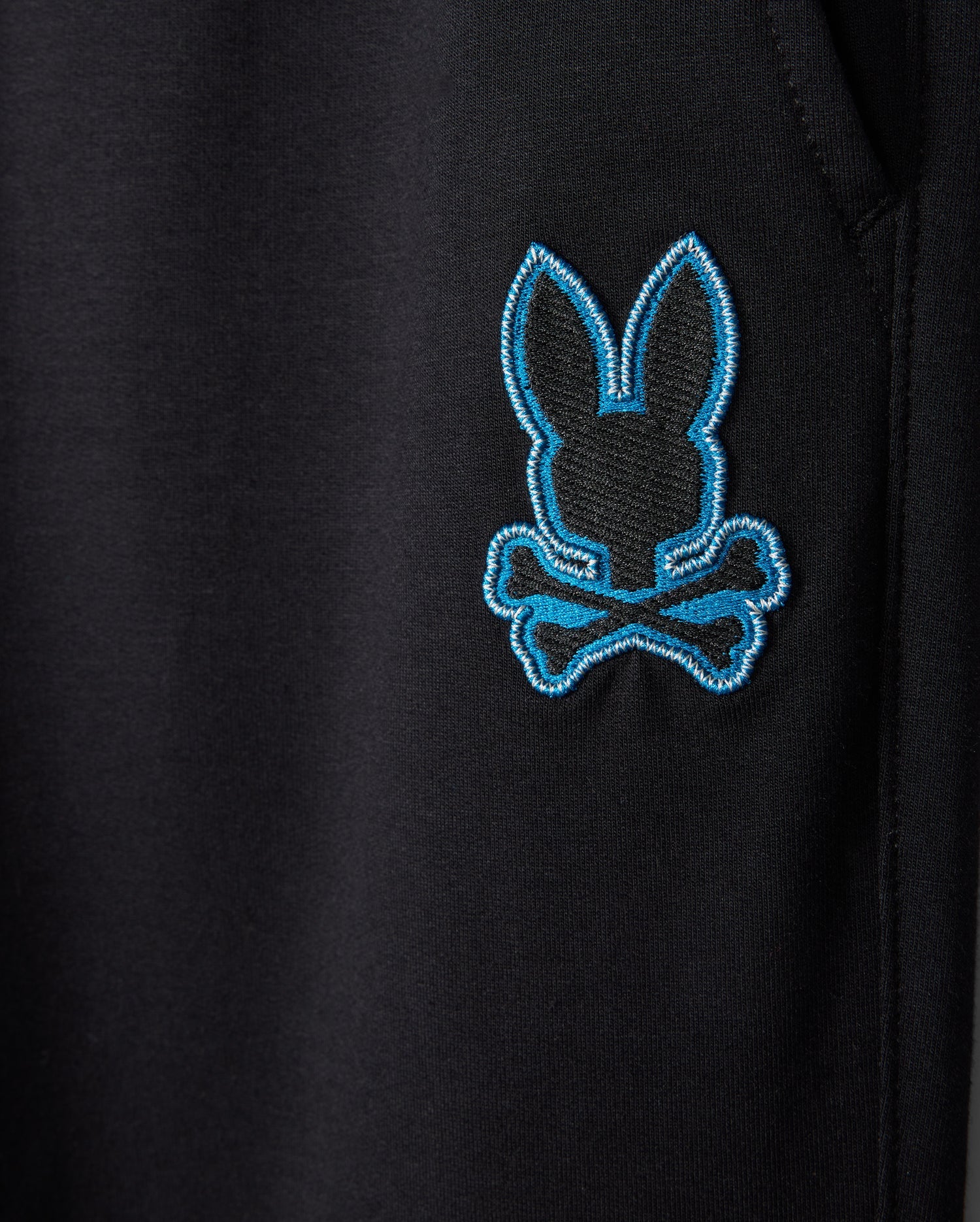 Close-up of a black fabric with an embroidered patch featuring a blue outline of the Psycho Bunny logo—a bunny head with crossbones underneath. The pocket seam is visible on the right side, adding character to casual wear. It's perfect for pairing with the breathable KIDS WALTER LIGHTWEIGHT SWEATPANT - B0P860D200 by Psycho Bunny.