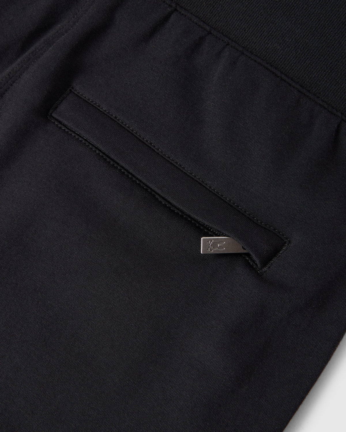 Close-up of a Psycho Bunny KIDS WALTER LIGHTWEIGHT SWEATPANT - B0P860D200 in black, featuring a pocket with a vertical opening and a silver metal tag attached to the edge. The fabric appears dense yet breathable, with neat and precise stitching that makes it perfect for casual wear.