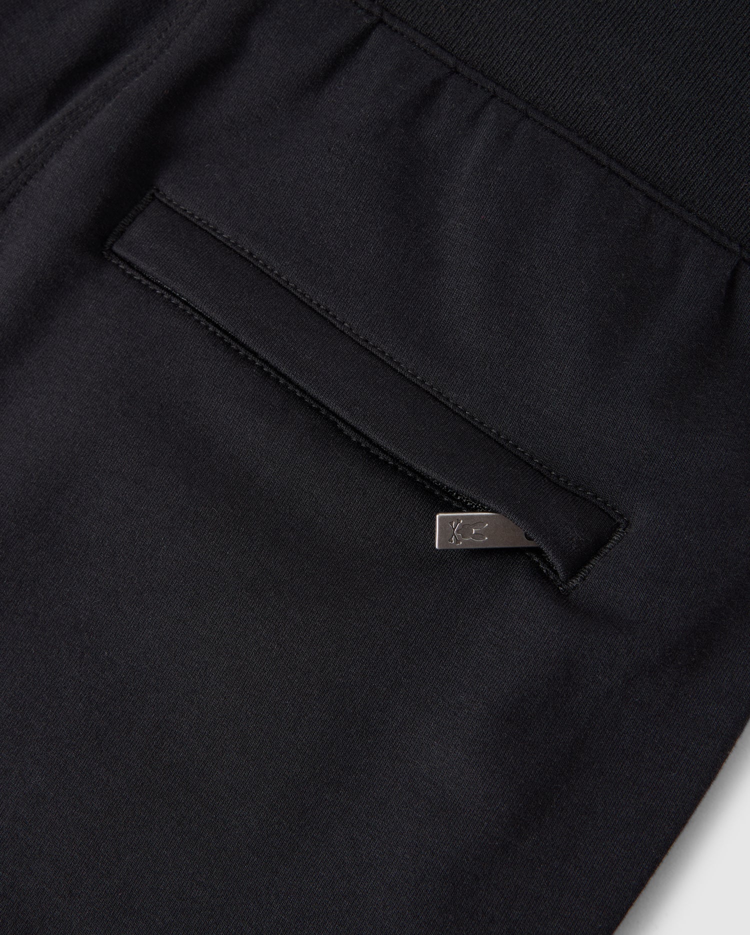 Close-up of a Psycho Bunny KIDS WALTER LIGHTWEIGHT SWEATPANT - B0P860D200 in black, featuring a pocket with a vertical opening and a silver metal tag attached to the edge. The fabric appears dense yet breathable, with neat and precise stitching that makes it perfect for casual wear.