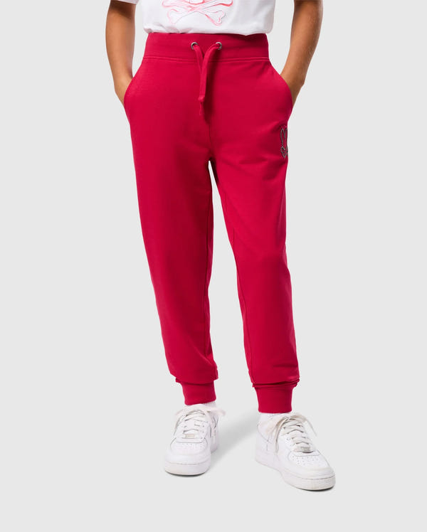 A person is wearing the bright red KIDS WALTER LIGHTWEIGHT SWEATPANT by Psycho Bunny, featuring drawstrings and pockets, paired with white sneakers. The background is plain gray, and only the lower half of the body is visible, highlighting the casual wear pants and shoes.