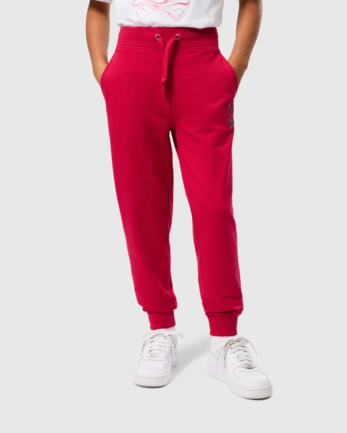 A person is wearing the bright red KIDS WALTER LIGHTWEIGHT SWEATPANT by Psycho Bunny, featuring drawstrings and pockets, paired with white sneakers. The background is plain gray, and only the lower half of the body is visible, highlighting the casual wear pants and shoes.