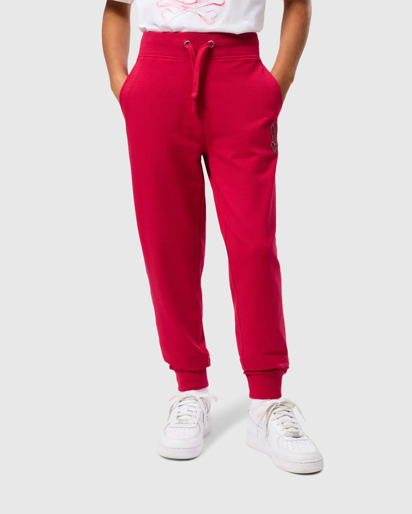 A person is wearing the bright red KIDS WALTER LIGHTWEIGHT SWEATPANT by Psycho Bunny, featuring drawstrings and pockets, paired with white sneakers. The background is plain gray, and only the lower half of the body is visible, highlighting the casual wear pants and shoes.