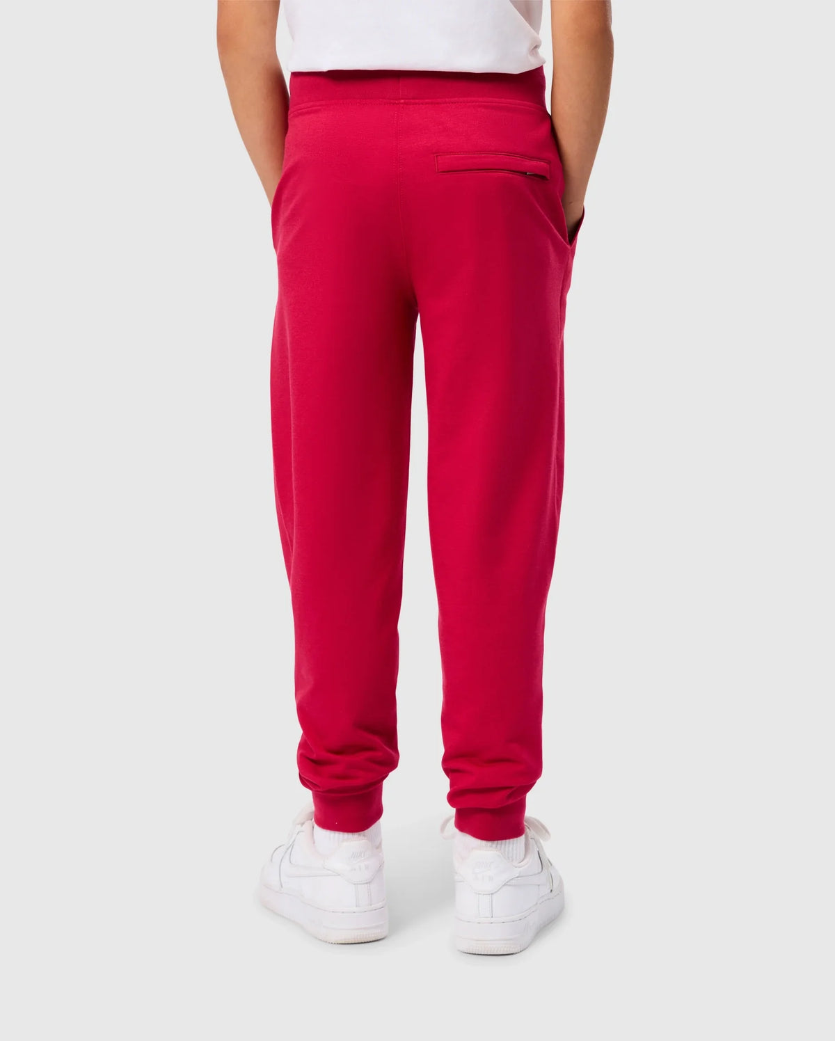 A child is seen from behind wearing the KIDS WALTER LIGHTWEIGHT SWEATPANT - B0P860D200 in a vibrant red color by Psycho Bunny, paired with a white shirt. The sweatpants feature two side pockets and one back pocket. White sneakers complete the outfit. The breathable fit of this casual wear contrasts nicely with the plain light gray background.