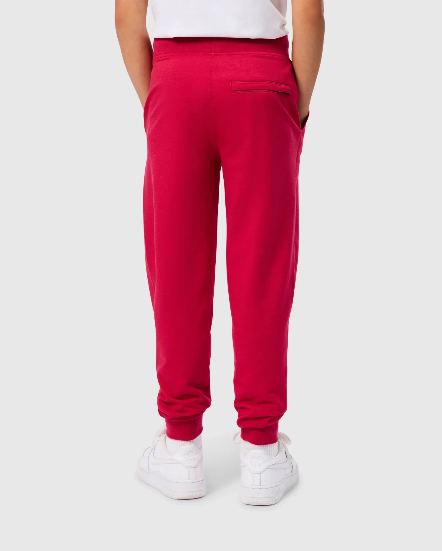 A child is seen from behind wearing the KIDS WALTER LIGHTWEIGHT SWEATPANT - B0P860D200 in a vibrant red color by Psycho Bunny, paired with a white shirt. The sweatpants feature two side pockets and one back pocket. White sneakers complete the outfit. The breathable fit of this casual wear contrasts nicely with the plain light gray background.
