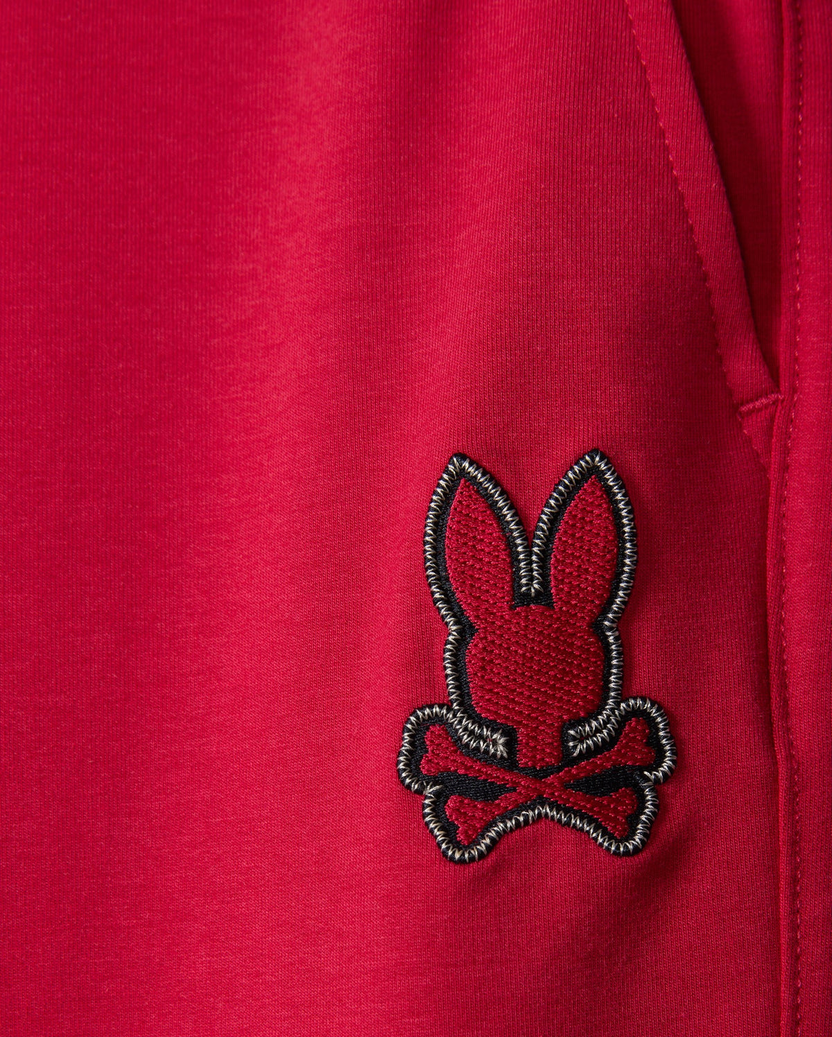 Close-up of the fabric from Psycho Bunny's KIDS WALTER LIGHTWEIGHT SWEATPANT (B0P860D200) in red, featuring a black and white embroidered patch with a bunny head and crossbones underneath. The lightweight sweatpant fabric showcases a visible seam and part of a pocket on the right side of the image, ideal for casual wear.