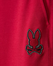 Close-up of the fabric from Psycho Bunny's KIDS WALTER LIGHTWEIGHT SWEATPANT (B0P860D200) in red, featuring a black and white embroidered patch with a bunny head and crossbones underneath. The lightweight sweatpant fabric showcases a visible seam and part of a pocket on the right side of the image, ideal for casual wear.