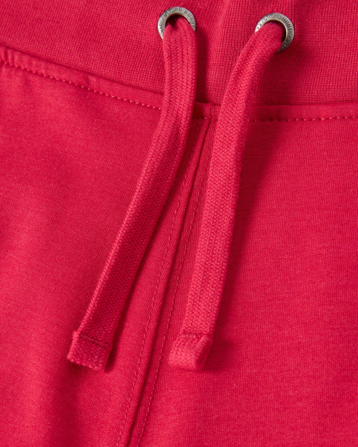 Close-up of the KIDS WALTER LIGHTWEIGHT SWEATPANT - B0P860D200 by Psycho Bunny in red, featuring drawstrings. The fabric appears soft and slightly textured, with two metal grommets through which the drawstrings are threaded. The seam is visible, indicating sturdy construction and ensuring a breathable fit ideal for casual wear.