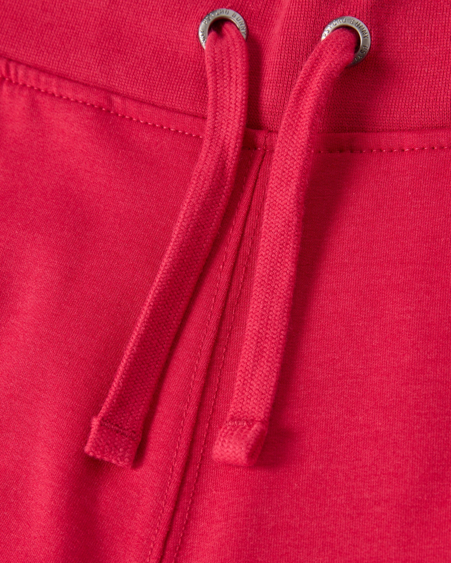 Close-up of the KIDS WALTER LIGHTWEIGHT SWEATPANT - B0P860D200 by Psycho Bunny in red, featuring drawstrings. The fabric appears soft and slightly textured, with two metal grommets through which the drawstrings are threaded. The seam is visible, indicating sturdy construction and ensuring a breathable fit ideal for casual wear.