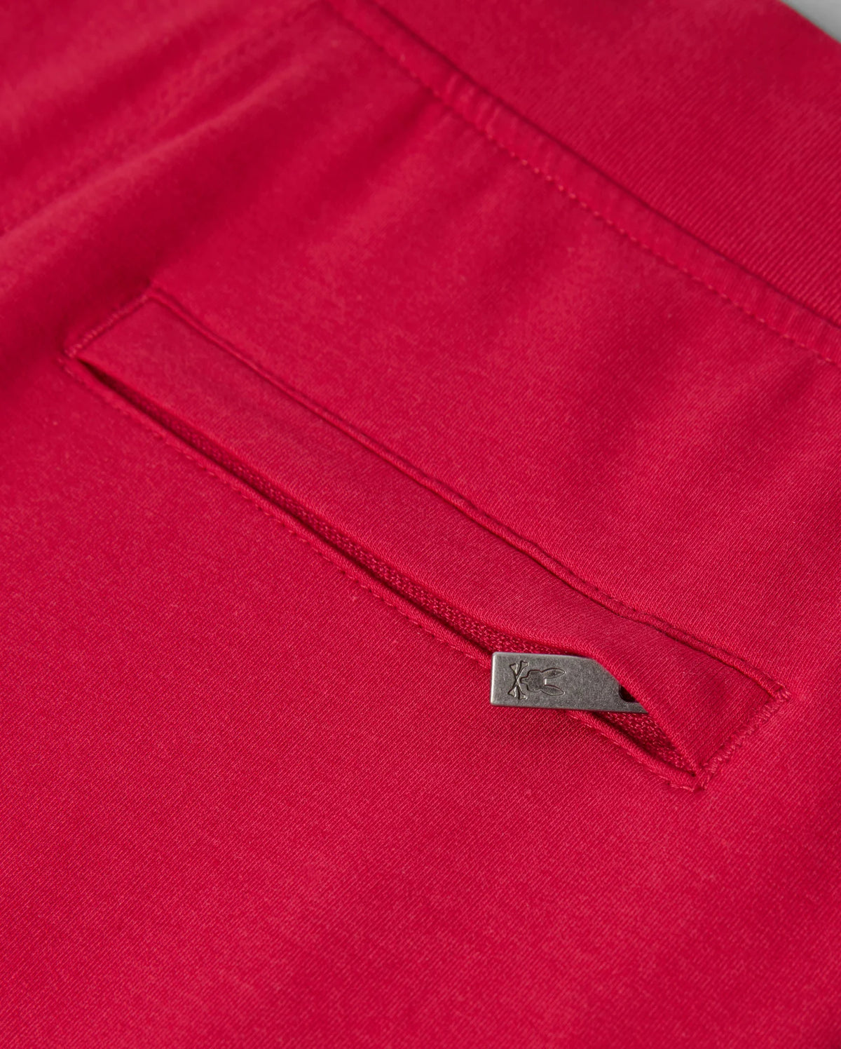 Close-up of the KIDS WALTER LIGHTWEIGHT SWEATPANT - B0P860D200 by Psycho Bunny in red, showcasing a seam with a concealed pocket. The pocket is adorned with a small metallic tab on its edge, featuring an engraved logo or symbol. This lightweight sweatpant fabric looks smooth and high-quality, ideal for casual wear with its breathable fit.