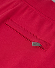 Close-up of the KIDS WALTER LIGHTWEIGHT SWEATPANT - B0P860D200 by Psycho Bunny in red, showcasing a seam with a concealed pocket. The pocket is adorned with a small metallic tab on its edge, featuring an engraved logo or symbol. This lightweight sweatpant fabric looks smooth and high-quality, ideal for casual wear with its breathable fit.