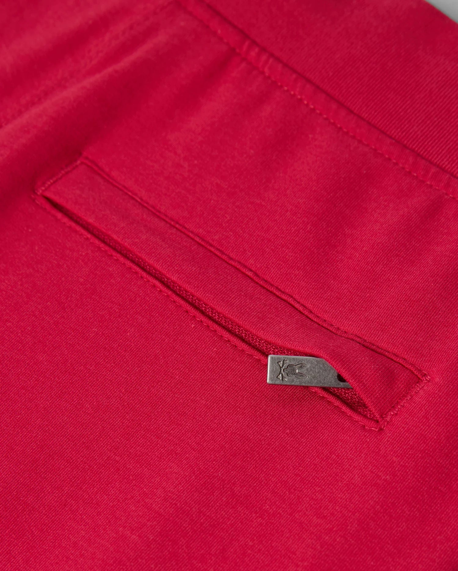 Close-up of the KIDS WALTER LIGHTWEIGHT SWEATPANT - B0P860D200 by Psycho Bunny in red, showcasing a seam with a concealed pocket. The pocket is adorned with a small metallic tab on its edge, featuring an engraved logo or symbol. This lightweight sweatpant fabric looks smooth and high-quality, ideal for casual wear with its breathable fit.