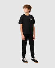 A young person with curly hair stands looking to the side, wearing a black T-shirt and Psycho Bunny's KIDS MONROE TWILL PATCH FLEECE SWEATPANTS. They complete their look with black sneakers featuring white soles, all set against a plain gray background.