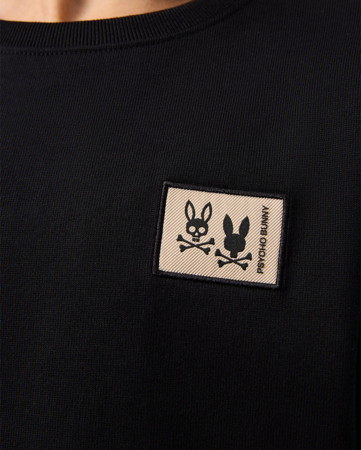 A black shirt showcases a small rectangular patch on the chest with a stylized bunny head and crossbones, beside "Psycho Bunny," crafted with luxury finishes. Pair it effortlessly with the KIDS MONROE TWILL PATCH FLEECE SWEATPANT - B0P929D200 for an elegant touch.