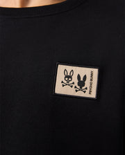 A black shirt showcases a small rectangular patch on the chest with a stylized bunny head and crossbones, beside "Psycho Bunny," crafted with luxury finishes. Pair it effortlessly with the KIDS MONROE TWILL PATCH FLEECE SWEATPANT - B0P929D200 for an elegant touch.