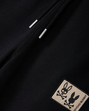 A close-up of the KIDS MONROE TWILL PATCH FLEECE SWEATPANT in black by Psycho Bunny features a zipper with metal pull tabs and luxury finishes, plus a bunny patch showcasing a stylized bunny skull with crossed bones and the text "Psycho Bunny.