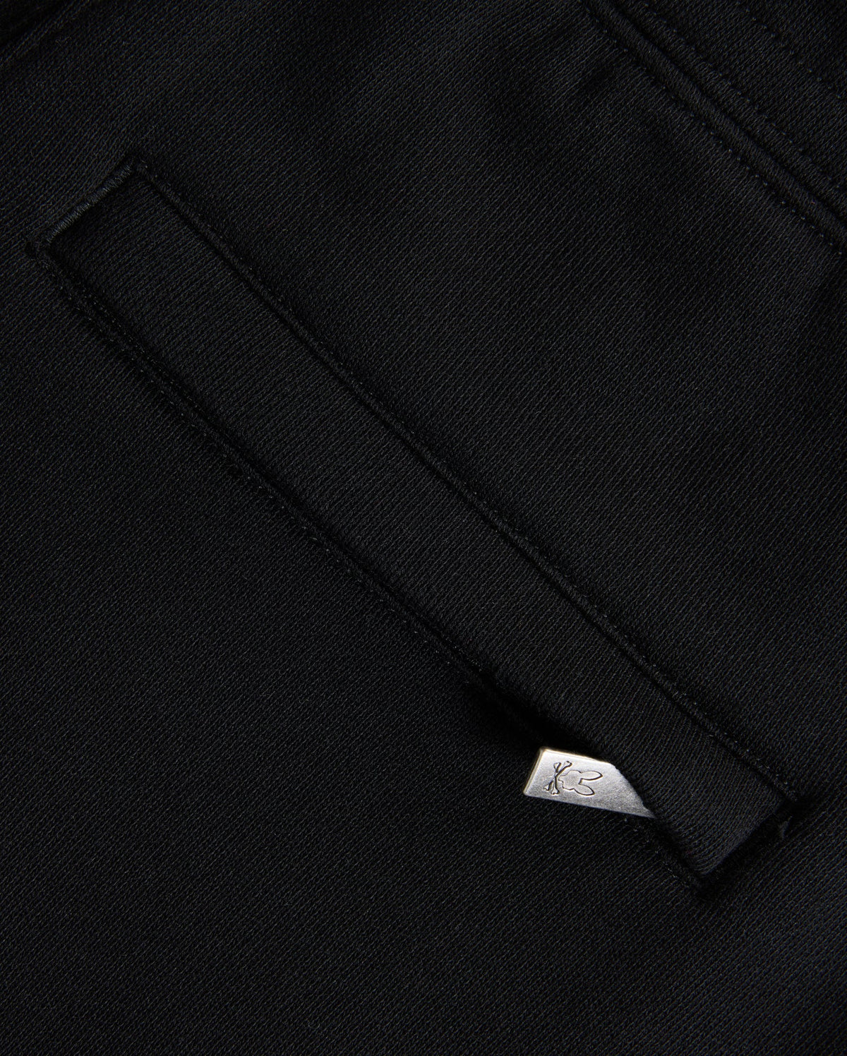 A close-up of black Psycho Bunny KIDS MONROE TWILL PATCH FLEECE SWEATPANTS showcases a welt pocket with a white tag featuring the logo. The slightly textured fabric and clean stitching evoke premium craftsmanship.