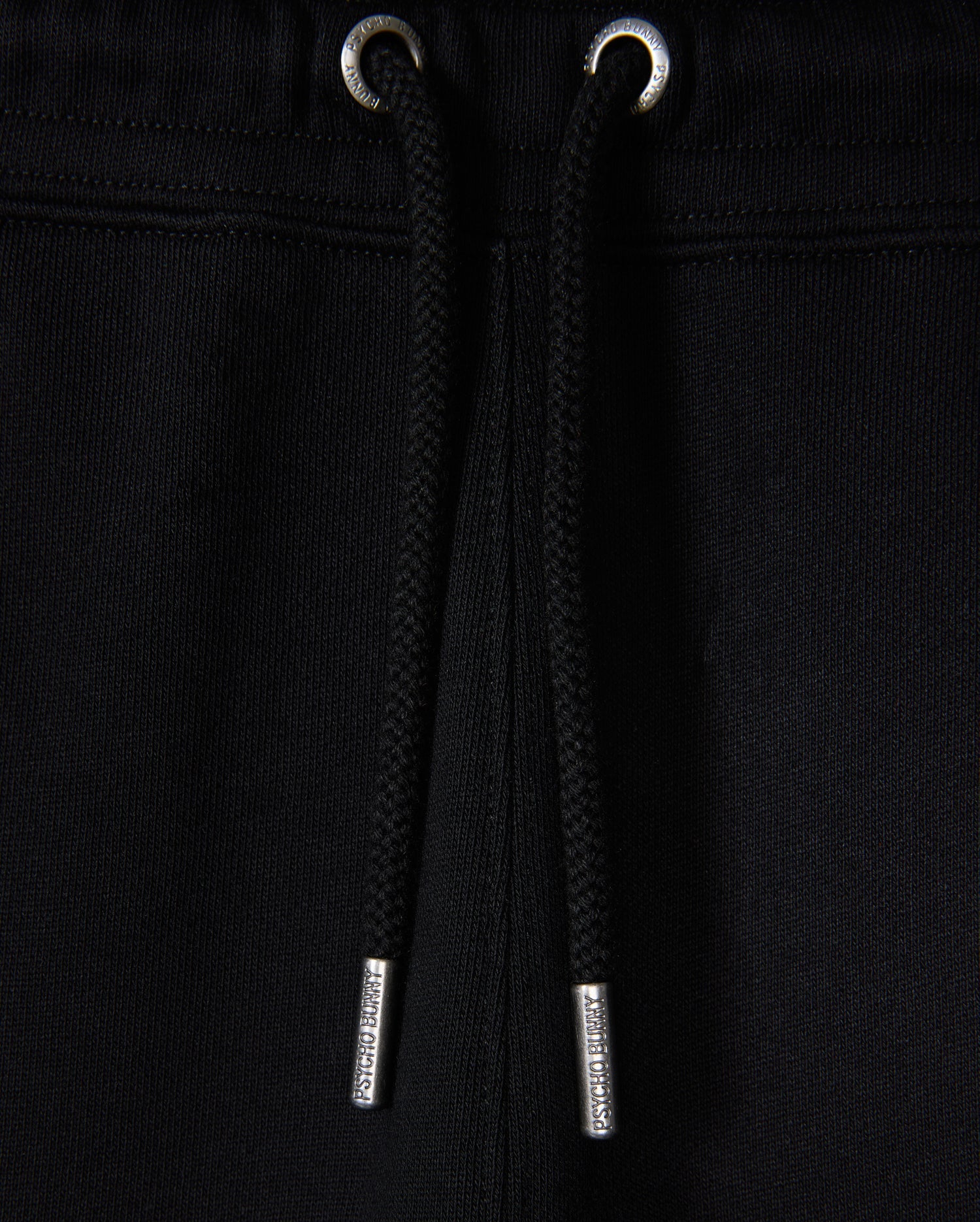 Close-up of black Psycho Bunny KIDS MONROE TWILL PATCH FLEECE SWEATPANT with luxury finishes, including drawstrings with metal tips threaded through metal eyelets at the waistband against a black fabric backdrop.