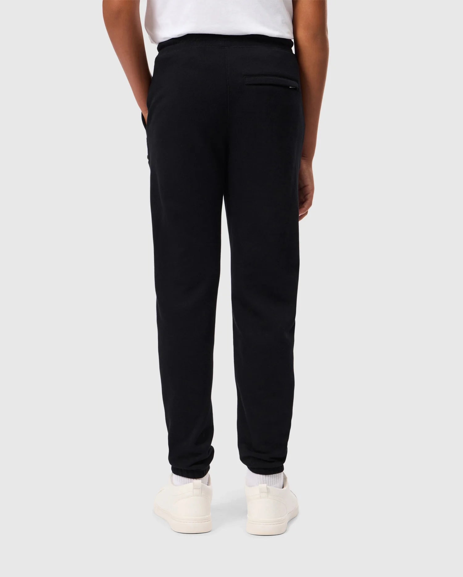 A person dressed in the Psycho Bunny's KIDS PACIFIC CHENILLE JOGGER stands with their back to the camera, wearing a white shirt and matching white sneakers. They casually tuck their hands into their pockets against a plain, light gray background.
