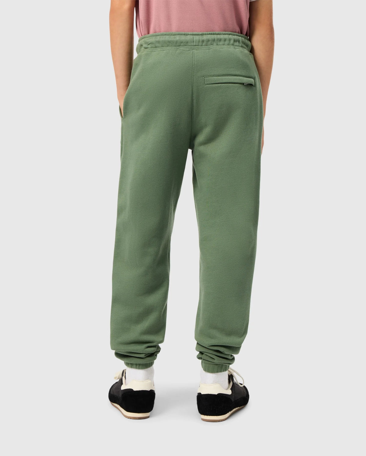 Wearing the KIDS PACIFIC CHENILLE JOGGER - B0P972D200 from Psycho Bunny, a person casually dressed in green faces away with hands in their pockets. These joggers feature a distinctive chenille logo patch and a right-side back pocket. They complete their look with black sneakers featuring white soles, perfectly paired with a pink shirt.