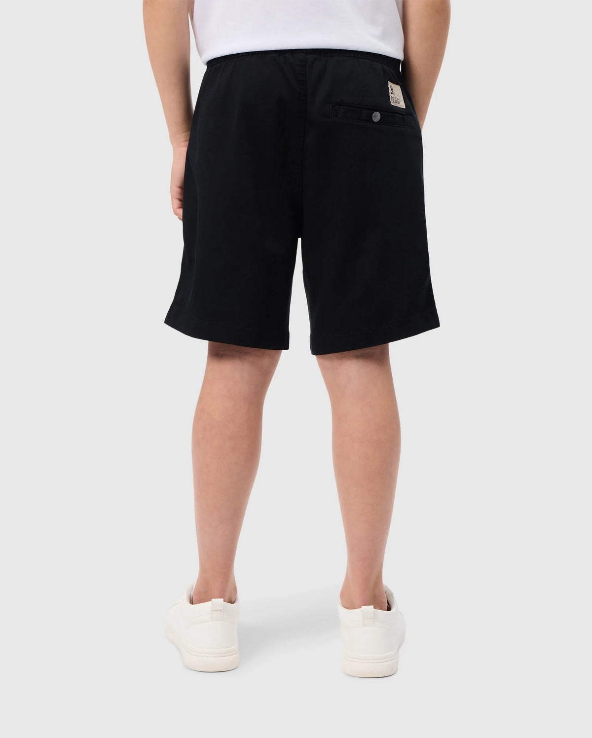 Rear view of a person wearing slim fit black shorts, a tucked-in white t-shirt, and white sneakers. The KIDS WILLIS STRETCH TENCEL SHORT - B0R239Y1WB from Psycho Bunny has a buttoned pocket on the right side with a beige tag above it and an embroidered Bunny logo. The person is standing against a plain grey background.