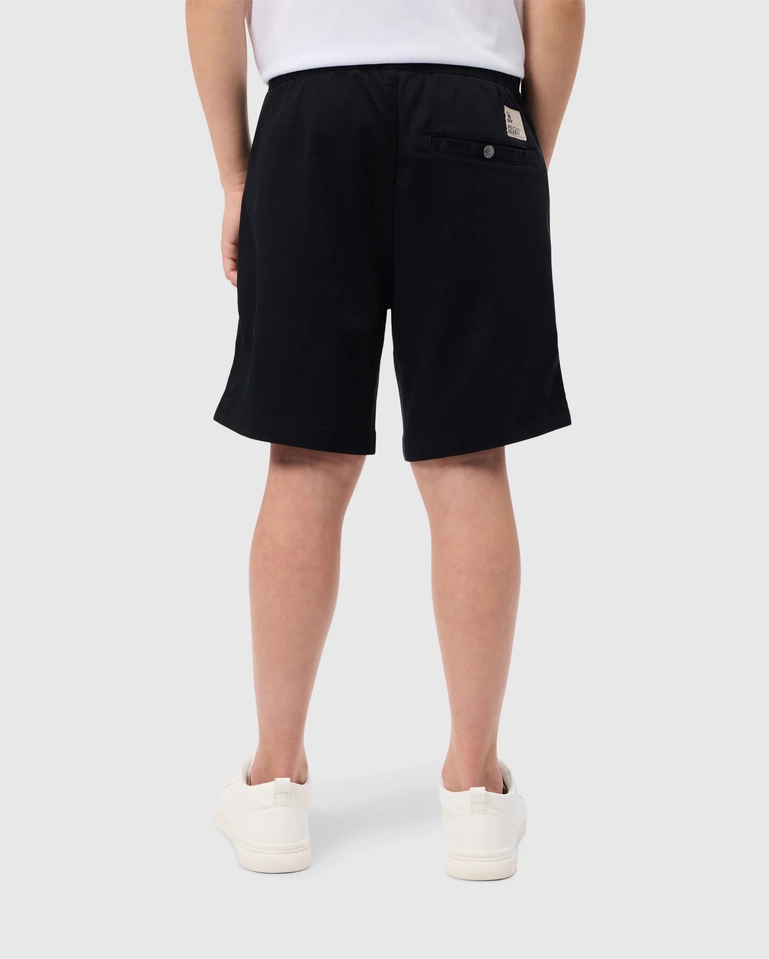 Rear view of a person wearing slim fit black shorts, a tucked-in white t-shirt, and white sneakers. The KIDS WILLIS STRETCH TENCEL SHORT - B0R239Y1WB from Psycho Bunny has a buttoned pocket on the right side with a beige tag above it and an embroidered Bunny logo. The person is standing against a plain grey background.