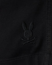 Close-up of black **KIDS WILLIS STRETCH TENCEL SHORT - B0R239Y1WB** fabric featuring an embroidered Bunny logo with ears upright, situated above a pair of crossed bones, reminiscent of a skull and crossbones motif. The slim fit fabric has visible stitching details. **Brand Name: Psycho Bunny**