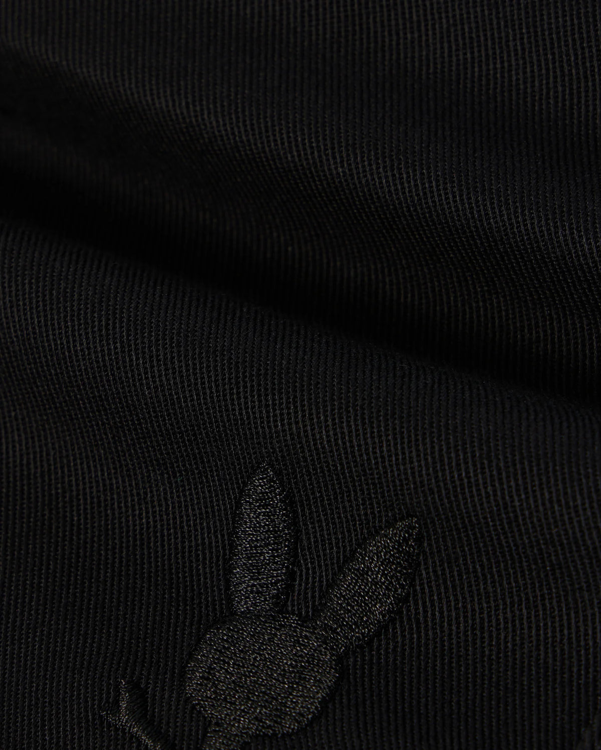 A close-up image of black fabric with a subtle embroidered Bunny logo featuring a rabbit's head with long ears on the lower portion. The texture of the KIDS WILLIS STRETCH TENCEL SHORT - B0R239Y1WB from Psycho Bunny is ribbed, and the embroidery is slightly raised, blending well with the dark color.