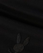 A close-up image of black fabric with a subtle embroidered Bunny logo featuring a rabbit's head with long ears on the lower portion. The texture of the KIDS WILLIS STRETCH TENCEL SHORT - B0R239Y1WB from Psycho Bunny is ribbed, and the embroidery is slightly raised, blending well with the dark color.