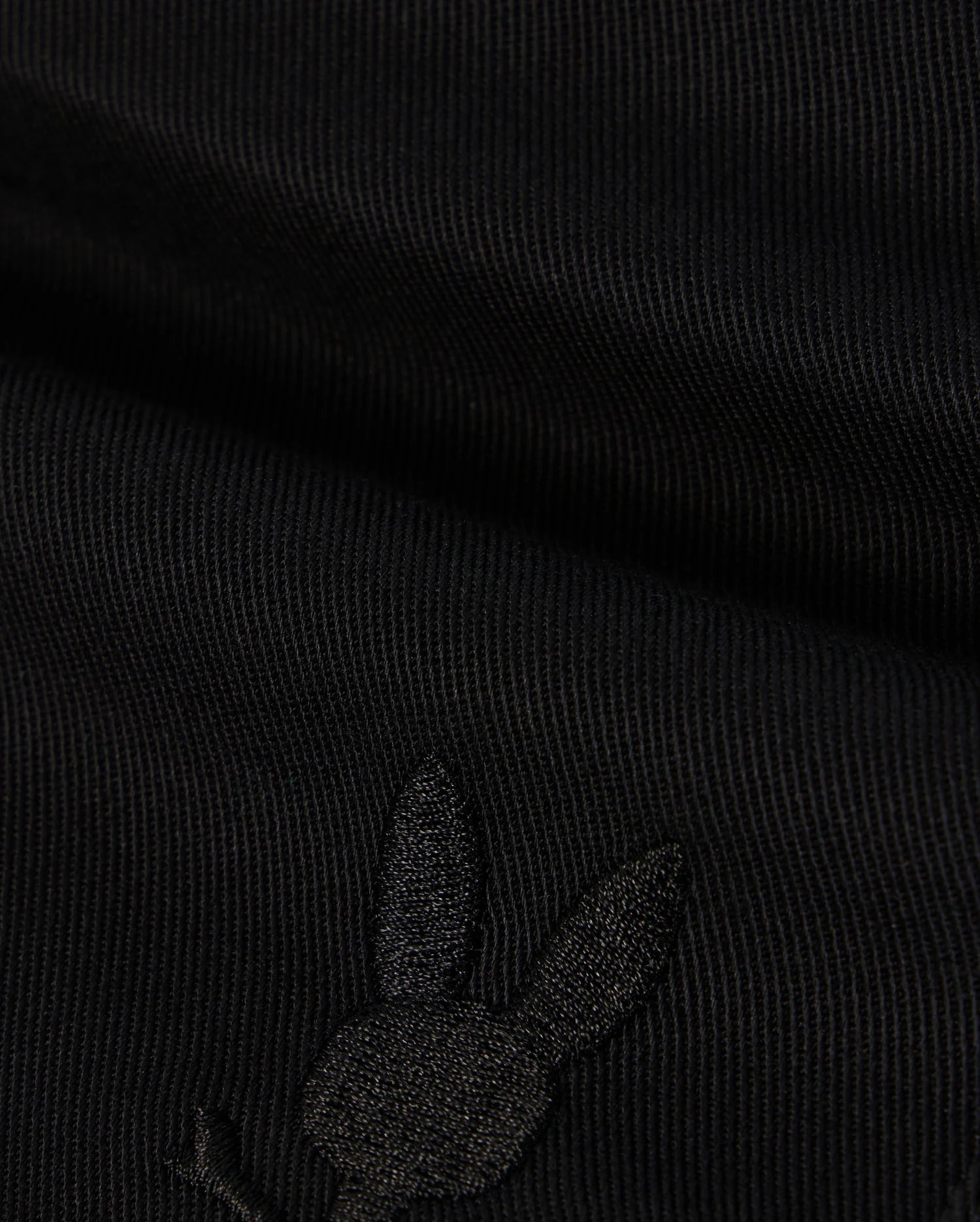 A close-up image of black fabric with a subtle embroidered Bunny logo featuring a rabbit's head with long ears on the lower portion. The texture of the KIDS WILLIS STRETCH TENCEL SHORT - B0R239Y1WB from Psycho Bunny is ribbed, and the embroidery is slightly raised, blending well with the dark color.