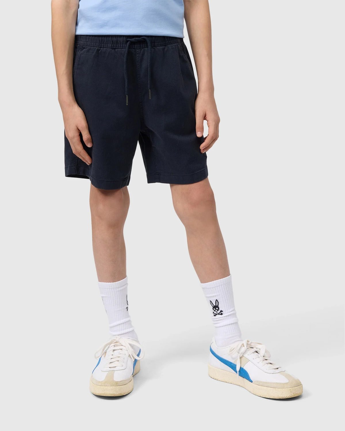 A person wearing Psycho Bunny navy blue elasticized waistband shorts, white socks, and white sneakers with blue accents, standing against a plain background. Only the lower half of the body is visible.
