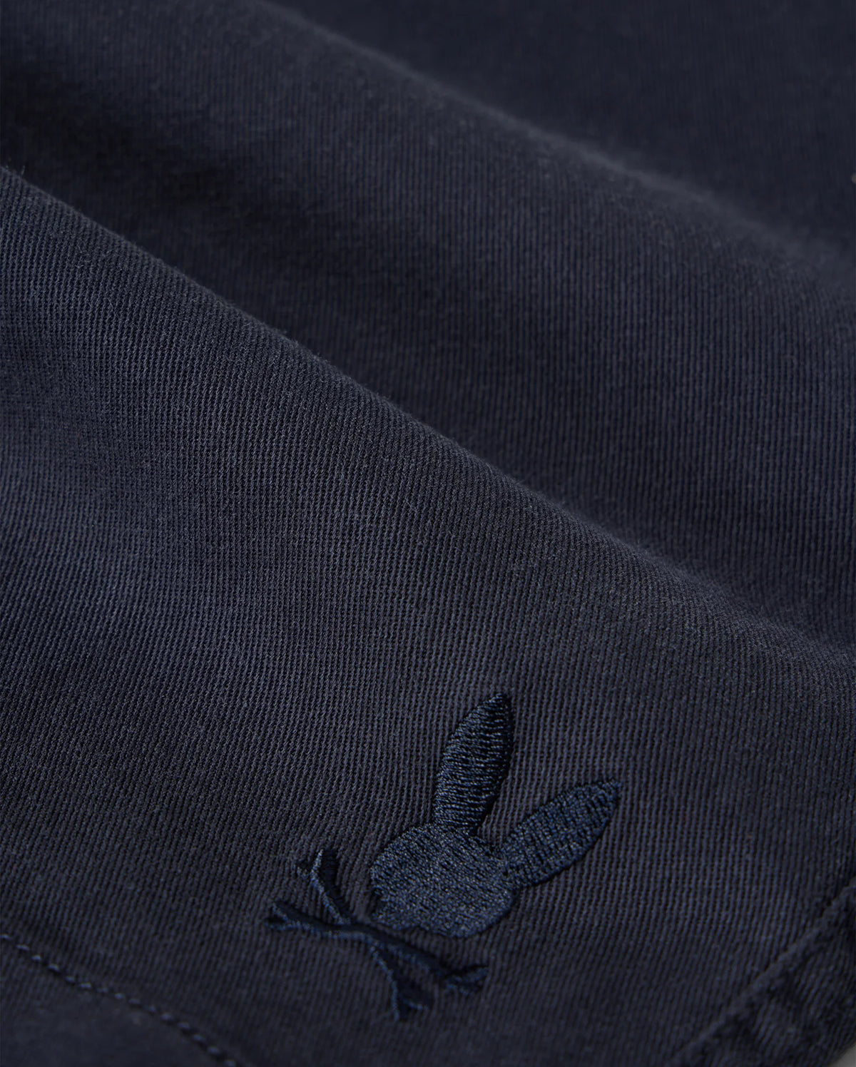 Close-up of Psycho Bunny kids' shorts made from dark KIDS WILLIS STRETCH TENCEL SHORT - B0R239Y1WB twill with a subtle, embroidered logo of a rabbit. The embroidery detail is in a slightly lighter shade of the same color as the fabric.