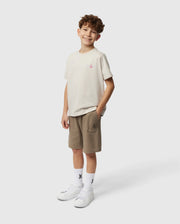 A smiling young boy with curly hair stands against a white background, wearing a lightweight Pima cotton beige T-shirt featuring a small chest design, white socks with graphics, and crisp white sneakers. He also has on Psycho Bunny's stylish KIDS STANFORD LIGHT PIQUE SWEATSHORT - B0R344B200 in khaki, his hands casually tucked into his pockets.
