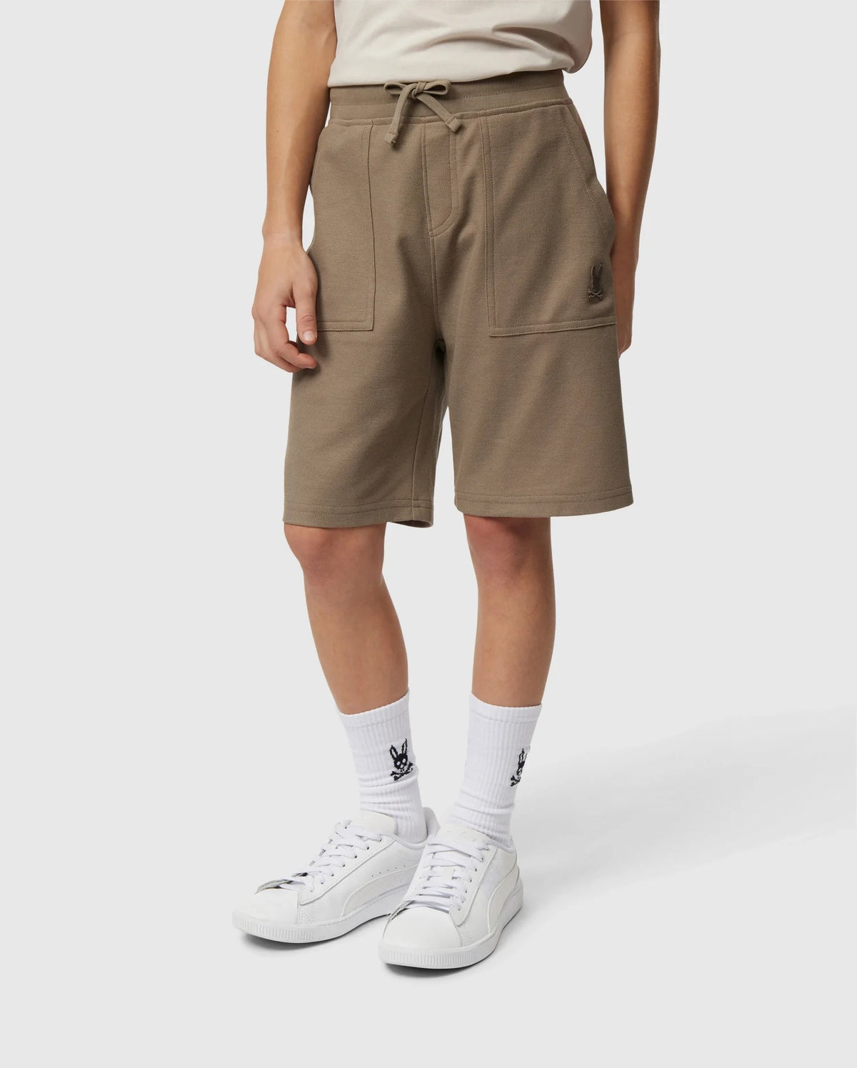 A person is wearing Psycho Bunny's KIDS STANFORD LIGHT PIQUE SWEATSHORT - B0R344B200, a stylish khaki knee-length garment made of lightweight Pima cotton, complete with front pockets and a drawstring waist. They have paired it with white socks that feature a black graphic design, and white sneakers. The individual stands against a plain white background.