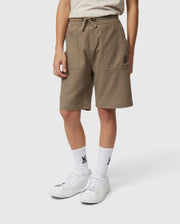 A person is wearing Psycho Bunny's KIDS STANFORD LIGHT PIQUE SWEATSHORT - B0R344B200, a stylish khaki knee-length garment made of lightweight Pima cotton, complete with front pockets and a drawstring waist. They have paired it with white socks that feature a black graphic design, and white sneakers. The individual stands against a plain white background.