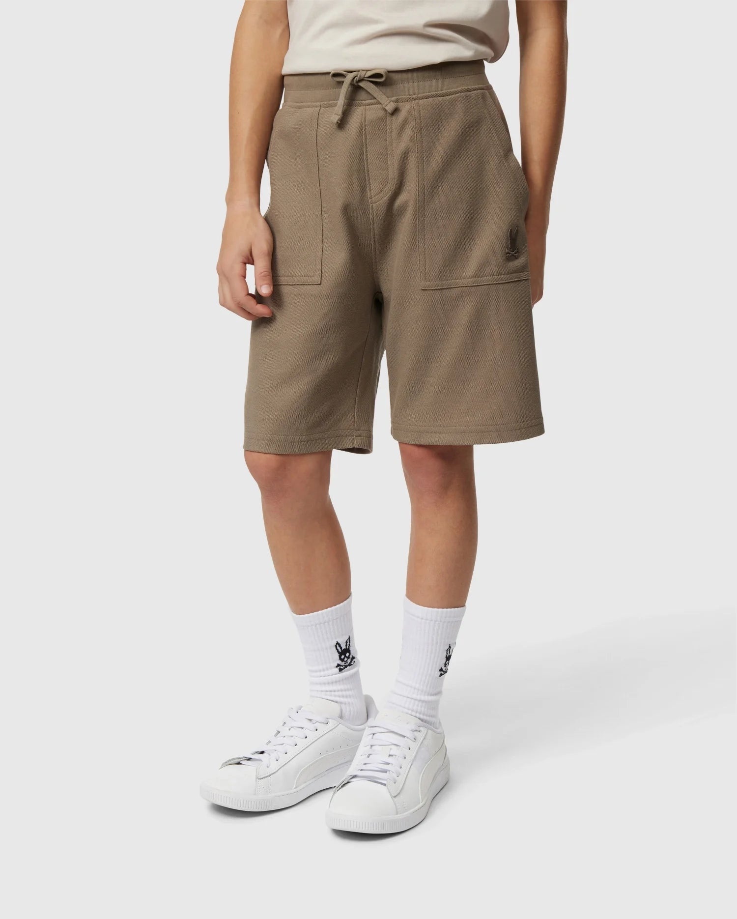 A person is wearing Psycho Bunny's KIDS STANFORD LIGHT PIQUE SWEATSHORT - B0R344B200, a stylish khaki knee-length garment made of lightweight Pima cotton, complete with front pockets and a drawstring waist. They have paired it with white socks that feature a black graphic design, and white sneakers. The individual stands against a plain white background.