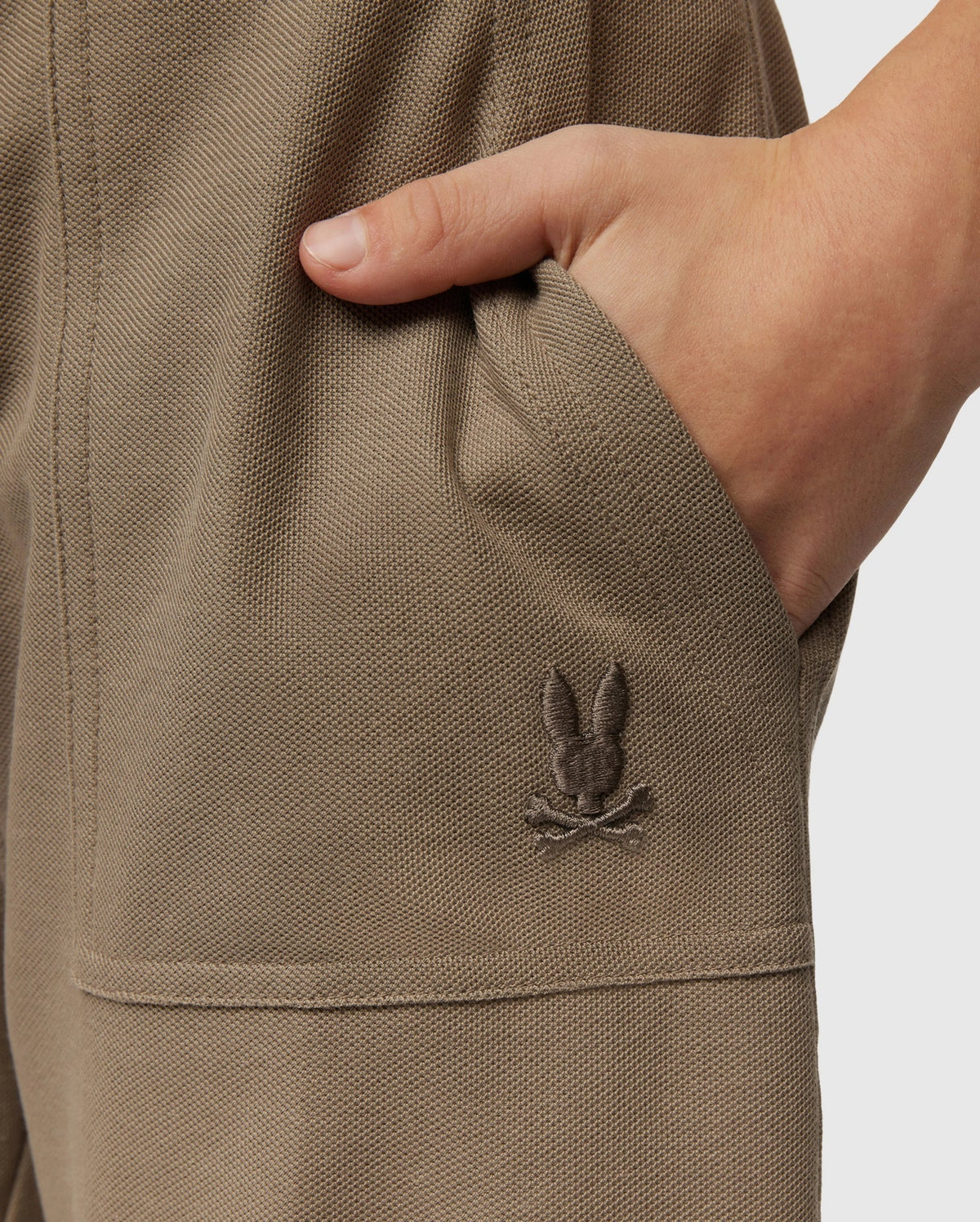Close-up of a child's hand tucked into the pocket of beige KIDS STANFORD LIGHT PIQUE SWEATSHORT - B0R344B200 by Psycho Bunny. Made from lightweight Pima cotton, these shorts feature an embroidered emblem of a bunny head with crossed bones beneath, located near the pocket. The fabric has a textured, slightly coarse appearance—truly a stylish wardrobe staple.