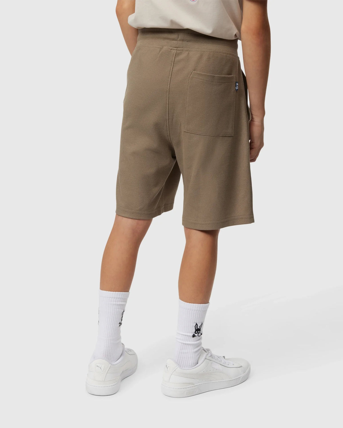 A person wearing a beige T-shirt and brown KIDS STANFORD LIGHT PIQUE SWEATSHORT - B0R344B200 from Psycho Bunny stands with their back to the camera. They are also wearing white socks and white Puma sneakers. The shorts, made from lightweight Pima cotton, feature a back pocket on the right side. The person is standing against a plain, white background.
