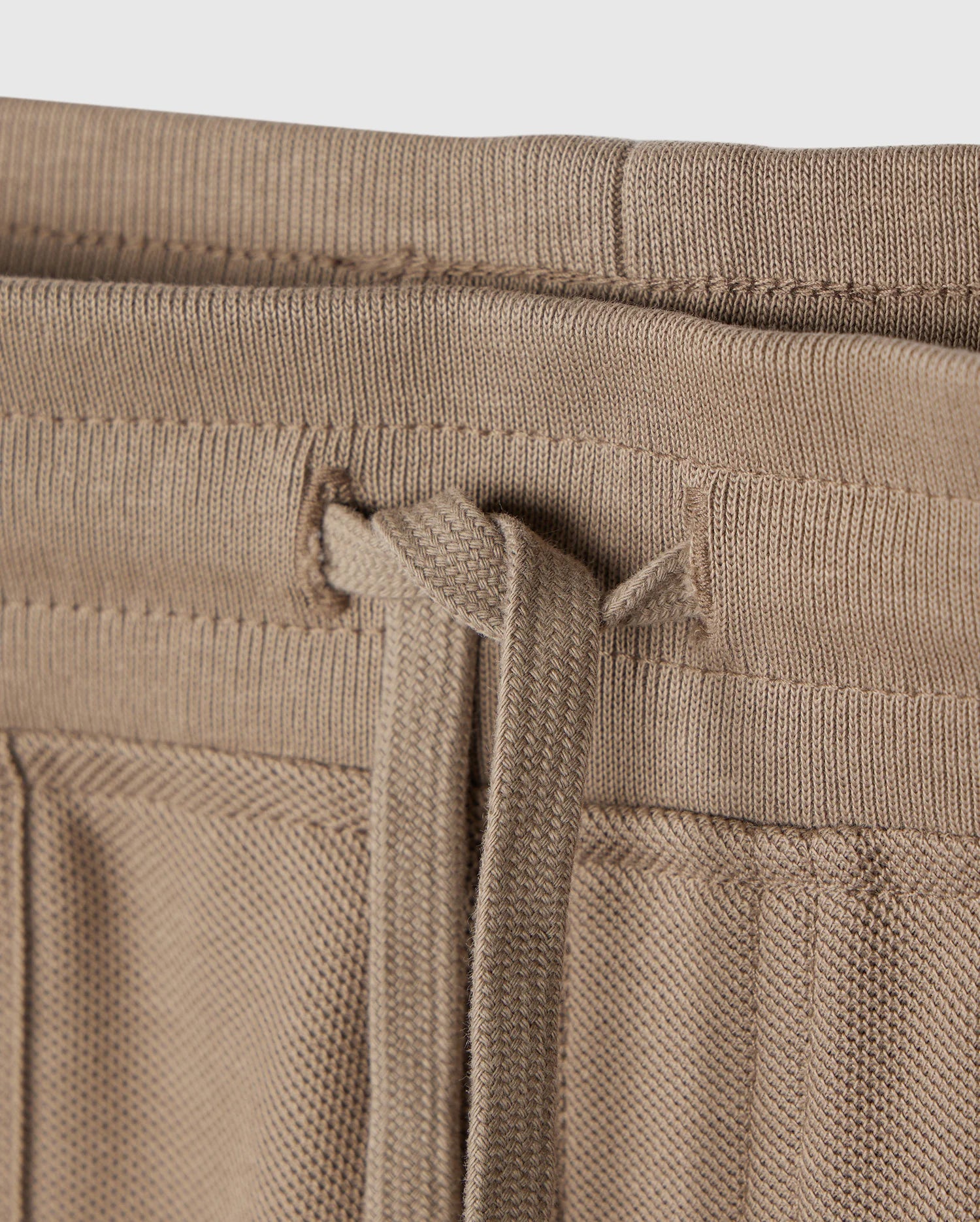 Close-up image showing the waistband of a pair of KIDS STANFORD LIGHT PIQUE SWEATSHORT - B0R344B200 from Psycho Bunny in beige, featuring a loosely tied drawstring threaded through sewn eyelets. The lightweight Pima cotton fabric has a textured appearance, and the stitching and detailing of this stylish wardrobe staple are clearly visible.