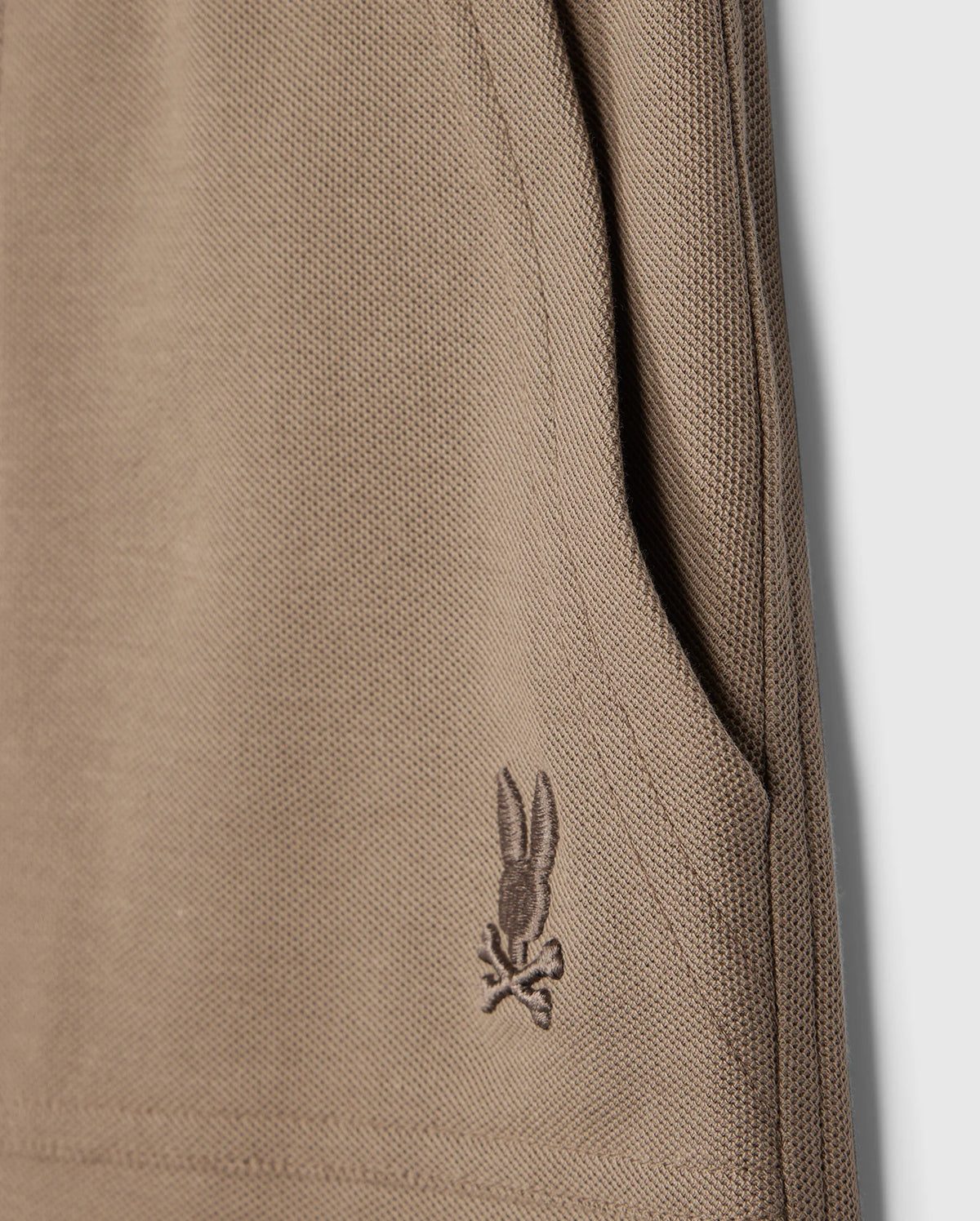 Close-up of the beige knit fabric from the KIDS STANFORD LIGHT PIQUE SWEATSHORT - B0R344B200 by Psycho Bunny, showing a pocket on the right side. Near the pocket's bottom edge, there is an embroidered design featuring a bunny with crossbones. The texture of the lightweight Pima cotton knit is clearly visible in the image.