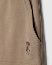 Close-up of the beige knit fabric from the KIDS STANFORD LIGHT PIQUE SWEATSHORT - B0R344B200 by Psycho Bunny, showing a pocket on the right side. Near the pocket's bottom edge, there is an embroidered design featuring a bunny with crossbones. The texture of the lightweight Pima cotton knit is clearly visible in the image.
