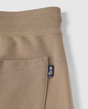 Close-up of a Kids Stanford Light Pique Sweatshort by Psycho Bunny in beige, zoomed in on the pocket and waistband. The fabric features a textured, waffle-like pattern. An attached navy tag with a white bunny logo is visible on the pocket's edge. Made from lightweight Pima cotton, this stylish wardrobe staple stands out against the simple, clean white background.