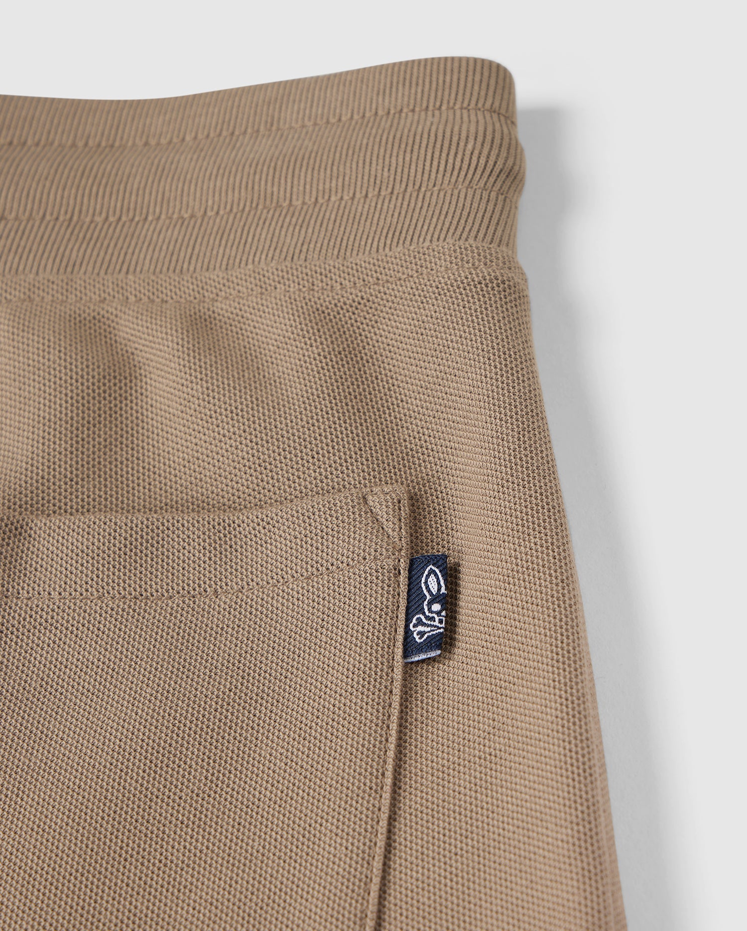 Close-up of a Kids Stanford Light Pique Sweatshort by Psycho Bunny in beige, zoomed in on the pocket and waistband. The fabric features a textured, waffle-like pattern. An attached navy tag with a white bunny logo is visible on the pocket's edge. Made from lightweight Pima cotton, this stylish wardrobe staple stands out against the simple, clean white background.