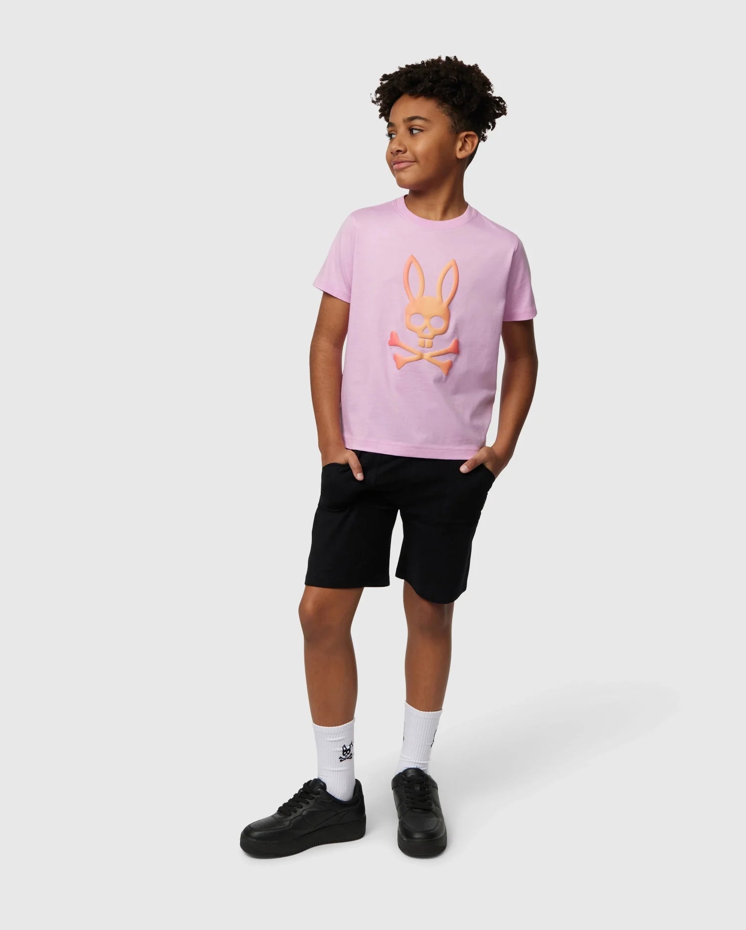 A young boy with curly hair stands with his hands in his pockets, wearing a pink Pima cotton piqué T-shirt featuring a bunny design and black Psycho Bunny KIDS STANFORD LIGHT PIQUE SWEATSHORT - B0R344B200. He is also wearing white socks and black shoes. He looks slightly to the side, smiling, against a plain white background.
