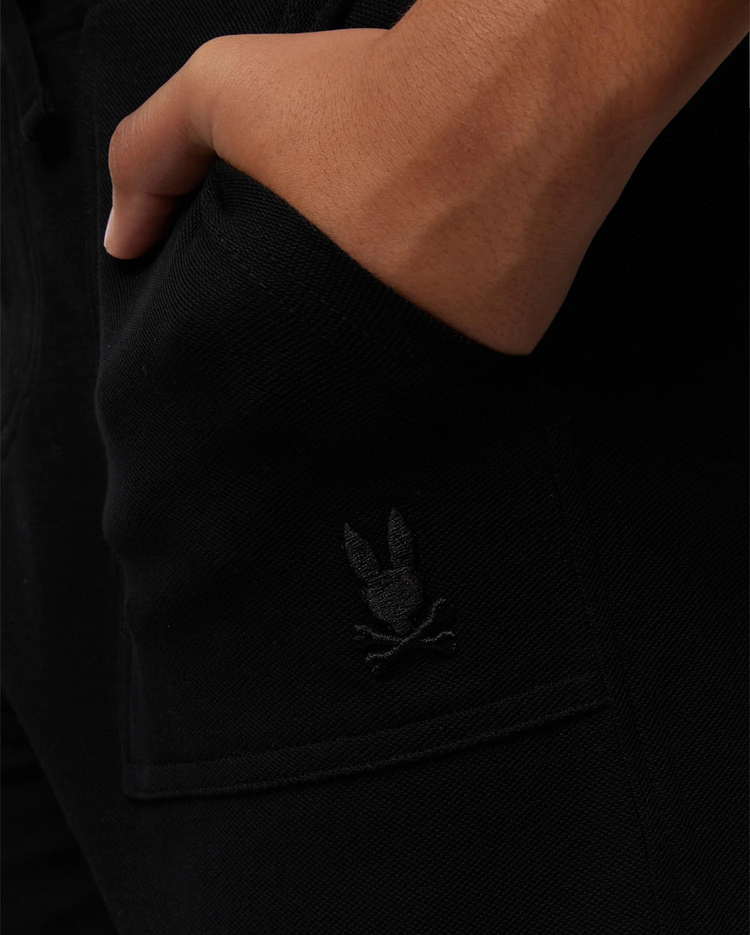 A close-up of a person’s hand resting in the pocket of black pants. The stylish wardrobe staple features an embroidered logo of a skull and crossbones with bunny ears on the pocket. The product is the KIDS STANFORD LIGHT PIQUE SWEATSHORT - B0R344B200 by Psycho Bunny.