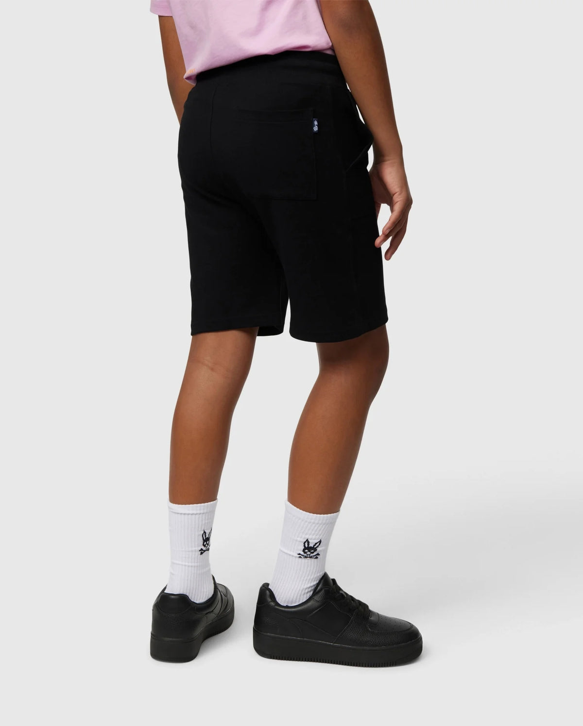 A person is standing with their back facing the camera, wearing a light pink Pima cotton piqué t-shirt, black KIDS STANFORD LIGHT PIQUE SWEATSHORT - B0R344B200 by Psycho Bunny, white socks featuring a small decorative logo, and black sneakers. The background is plain white.