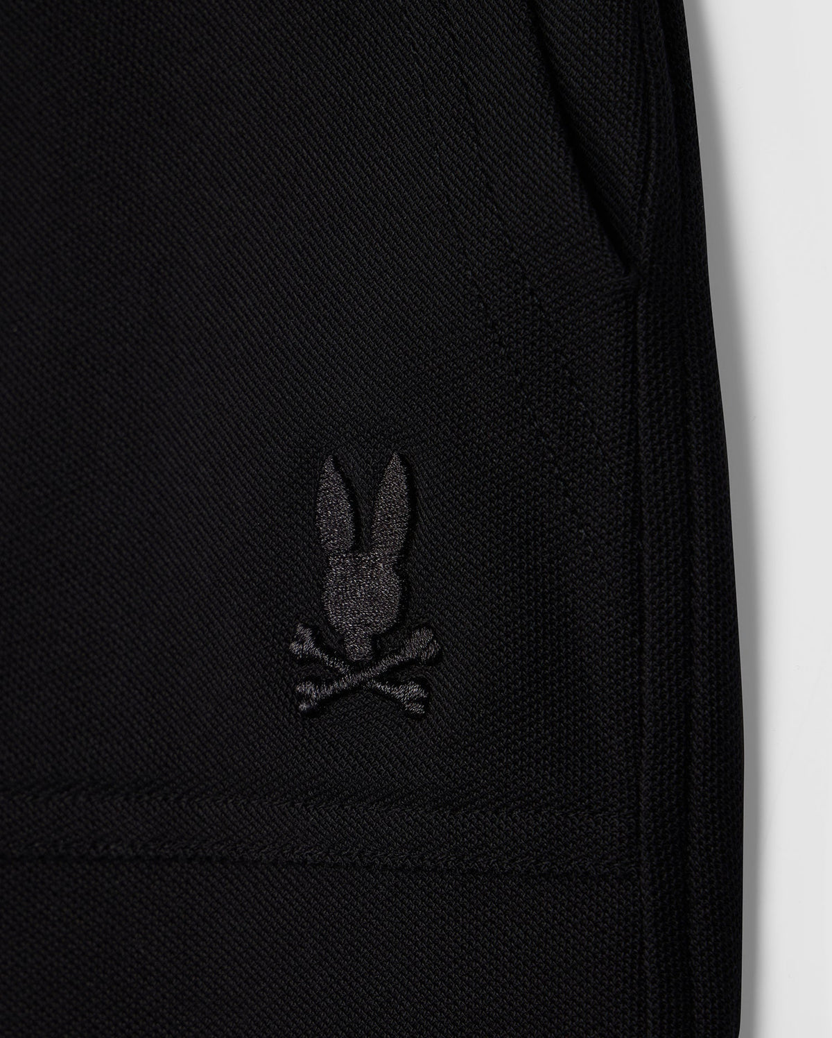 Close-up image of a black Pima cotton piqué fabric featuring an embroidered black logo of a rabbit head with crossed bones beneath it, resembling a skull and crossbones design. The image highlights the texture and details of the stitching on this stylish wardrobe staple, the KIDS STANFORD LIGHT PIQUE SWEATSHORT - B0R344B200 from Psycho Bunny.