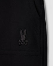 Close-up image of a black Pima cotton piqué fabric featuring an embroidered black logo of a rabbit head with crossed bones beneath it, resembling a skull and crossbones design. The image highlights the texture and details of the stitching on this stylish wardrobe staple, the KIDS STANFORD LIGHT PIQUE SWEATSHORT - B0R344B200 from Psycho Bunny.