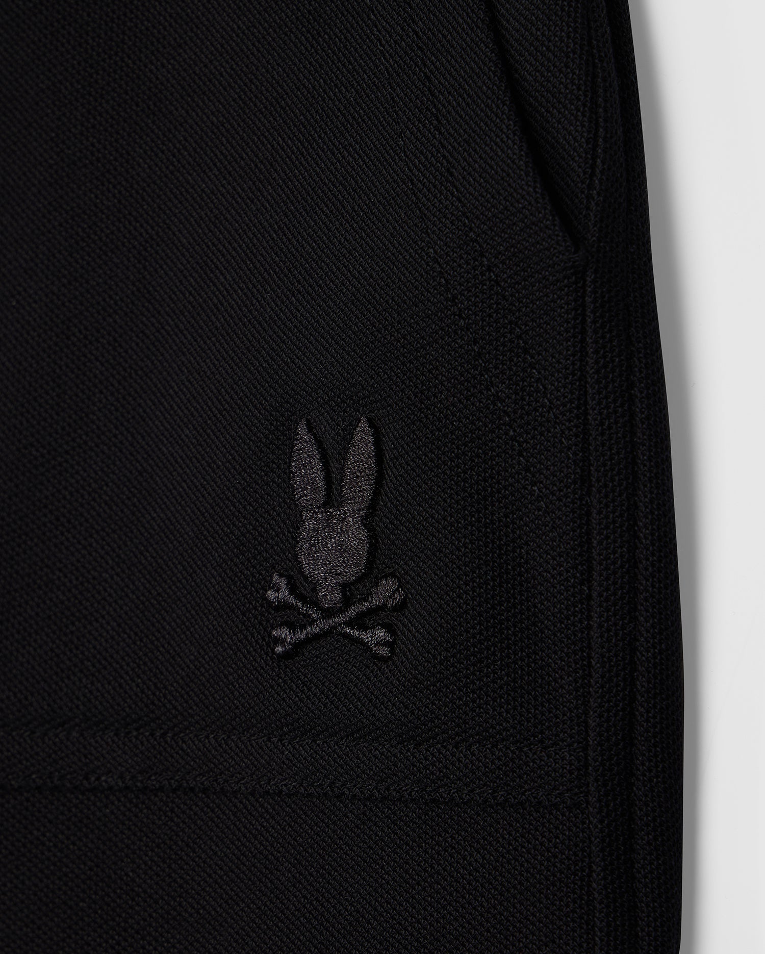 Close-up image of a black Pima cotton piqué fabric featuring an embroidered black logo of a rabbit head with crossed bones beneath it, resembling a skull and crossbones design. The image highlights the texture and details of the stitching on this stylish wardrobe staple, the KIDS STANFORD LIGHT PIQUE SWEATSHORT - B0R344B200 from Psycho Bunny.
